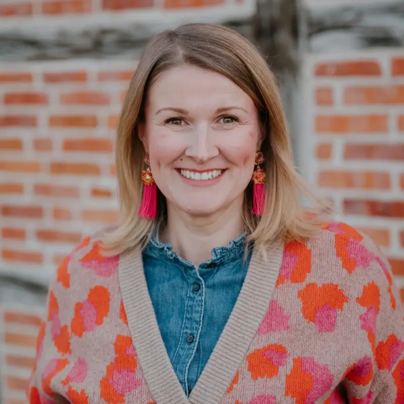 Anne Löwenstein lächelt in die Kamera und ist Inhaberin von Anne Löwenstein Immobilien. Im Hintergrund ist das rote Backsteinhaus das Maklerbüro.